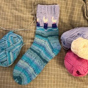 May include: A blue and purple striped knitted sock with a purple cuff featuring three white birds. The sock is lying on a brown fabric surface with balls of yarn in various colors.