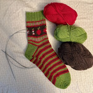 May include: A green, red, and brown striped sock with a brown, green, and pink apple design. Three balls of yarn are in the background: red, green, and brown.