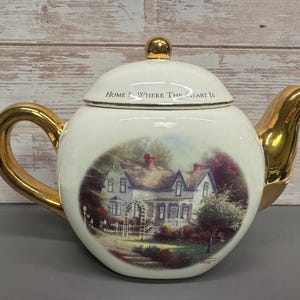 May include: A white ceramic teapot with a gold handle and spout. The teapot has a painting of a house with a garden on the front. The text "Home Is Where The Heart Is" is written on the lid.
