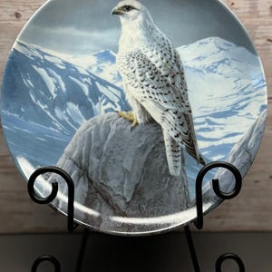 May include: Decorative plate featuring a white falcon perched on a rock, set against a backdrop of snow-capped mountains. The plate has a blue and white color scheme, and is displayed on a black metal stand. The plate's design is detailed and realistic.