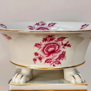 May include: A white porcelain bowl with a floral design in shades of pink and gold trim. The bowl is supported by four white paw feet. The bowl is approximately 12 inches in diameter.