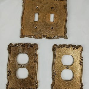 May include: Three gold-colored metal wall plates with a textured finish and ornate, scalloped edges. One plate features two rectangular toggle switch openings, while the other two plates each feature two circular duplex outlet openings.