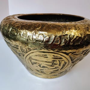 May include: A large, gold-colored brass planter with an intricate embossed design. The planter has a wide, round opening and a slightly flared base.