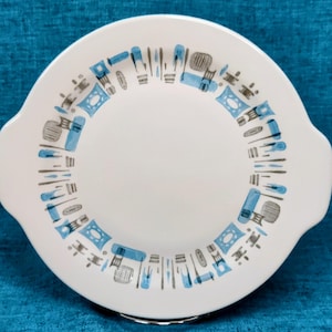 May include: White ceramic serving plate with two handles. The plate features a blue and gray geometric pattern around the rim. The design includes rectangular and oblong shapes. The plate is round and has a diameter of approximately 10 inches.