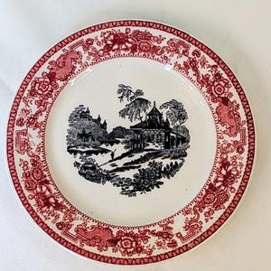 May include: A round ceramic plate with a white center and a red decorative border featuring floral patterns. The center of the plate displays a black and white transfer print illustration of a landscape with a building, trees, and a small figure.