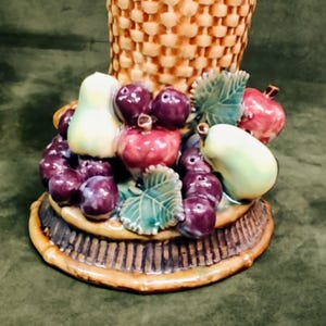 May include: A ceramic basket-shaped vase with a woven texture. The base is adorned with a colorful arrangement of ceramic fruits, including grapes, apples, and pears. The fruits are painted in shades of red, purple, and green, creating a decorative centerpiece.