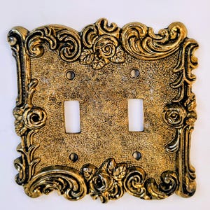 May include: A double light switch wall plate with a metallic gold finish and a textured surface. The rectangular plate features ornate raised scrollwork and rose floral motifs along the edges, with two vertical rectangular cutouts for switches.