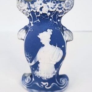 May include: A blue and white ceramic vase featuring a raised white relief of a figure holding a lyre. The vase has a flared rim decorated with floral motifs and a scalloped base. The central design depicts a person in profile with flowing hair.