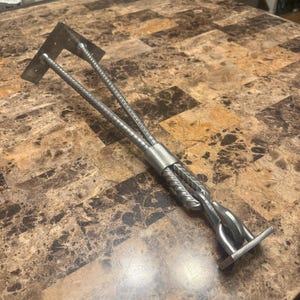 May include: A handcrafted metal tool with a unique design. It features two parallel, textured rods connected by a metal sleeve, with flat, rectangular ends. The tool is made of silver-colored metal and rests on a brown and tan patterned surface.