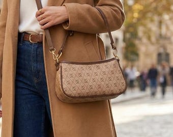 May include: A woman wearing a camel-colored long coat, white top, and dark blue jeans. She carries a brown shoulder bag with a repeating pattern. Brown leather boots complete the outfit. The scene is set on a cobblestone street.
