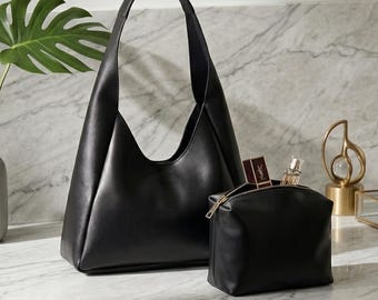 May include: A black leather shoulder bag and matching cosmetic bag sit on a marble countertop. The shoulder bag has a single handle and a triangular shape. The cosmetic bag is rectangular with a zipper. A vase with a green leaf and perfume bottles are also present.