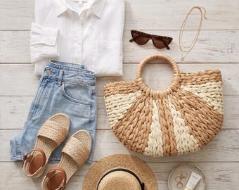 May include: A flat lay of summer fashion essentials. Includes a white button-down shirt, light-wash denim shorts, woven sandals, a straw tote bag, sunglasses, a sun hat, and gold jewelry. A small dish holds rings and sunscreen.