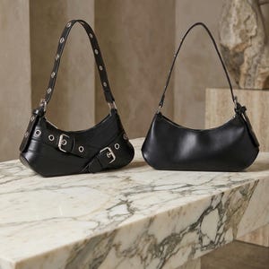 May include: Two black leather shoulder bags on a marble surface. One bag features silver buckles and grommets, while the other has a minimalist design. Both have curved silhouettes and thin straps, showcasing a modern aesthetic.