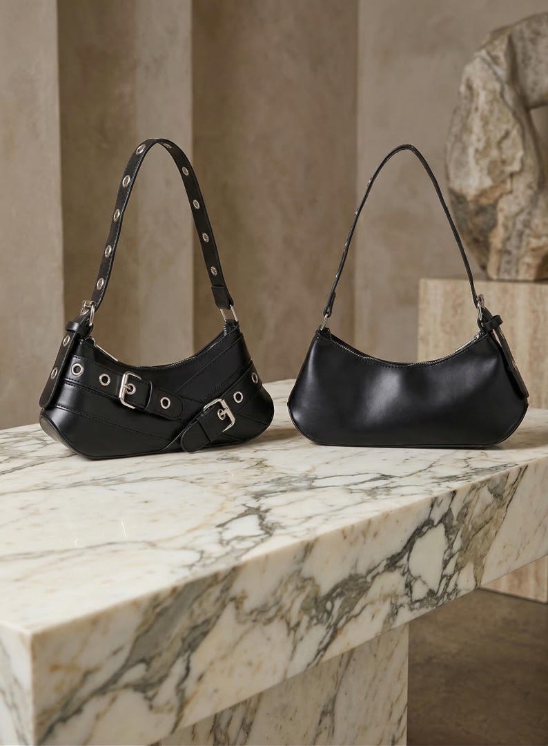 May include: Two black leather shoulder bags on a marble surface. One bag features silver buckles and grommets, while the other has a minimalist design. Both have curved silhouettes and thin straps, showcasing a modern aesthetic.