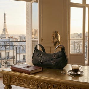 May include: A black leather shoulder bag with multiple buckles sits on a gold table, next to a leather-bound journal and a cup of coffee. The Eiffel Tower is visible through an open window in the background.