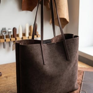 May include: A large, brown suede tote bag with long, thin handles. The bag is rectangular with a wide opening and sits on a wooden surface. The background includes leather hides and tools, suggesting a handcrafted item.