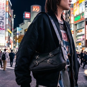 May include: A black leather shoulder bag with silver buckles is worn over a black jacket. The bag has a rectangular shape with three buckles on the front. The background shows a busy city street with illuminated signs and pedestrians.