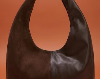 May include: A dark brown shoulder bag with a crescent shape. The bag is made of two different materials, with a suede-like texture on the left side and a smooth leather-like texture on the right side. The bag has a single strap.