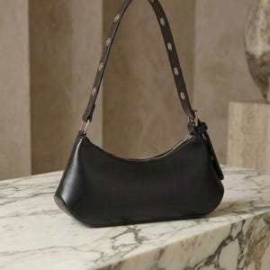 May include: Black leather shoulder bag with a curved shape and silver-tone hardware. The strap is adorned with small, round silver studs. The bag is displayed on a marble surface.