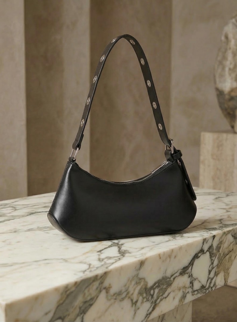 May include: Black leather shoulder bag with a curved shape and silver-tone hardware. The strap is adorned with small, round silver studs. The bag is displayed on a marble surface.