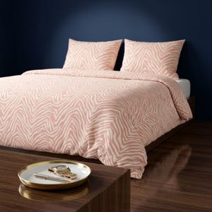 May include: A bedding set featuring a duvet cover and two matching pillowcases with a light pink and white zebra stripe pattern. In the foreground, a round white tray with a gold rim holds a silver pen and a gold-toned wristwatch.