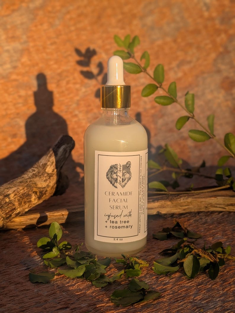May include: A clear glass bottle of Ceramide Facial Serum with a gold dropper top. The label features a wolf illustration and the text "infused with tea tree + rosemary". The bottle contains 3.4 oz of serum and is surrounded by greenery.