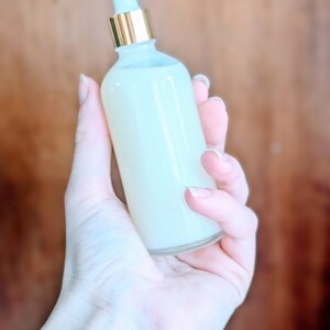 May include: A clear glass bottle filled with a creamy, pale yellow liquid. The bottle has a gold-colored collar and a light blue dropper. The bottle is held in a person's hand against a blurred brown background.