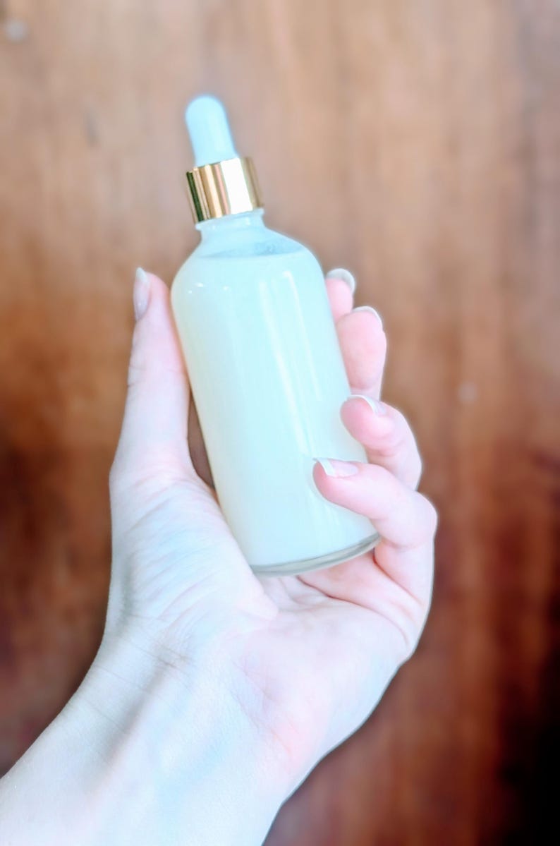 May include: A clear glass bottle filled with a creamy, pale yellow liquid. The bottle has a gold-colored collar and a light blue dropper. The bottle is held in a person's hand against a blurred brown background.