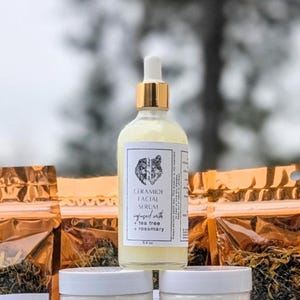 May include: A clear glass bottle of Ceramide Facial Serum with a gold dropper top. The label features a wolf design and text indicating it's infused with tea tree and rosemary. The bottle is 3.4 fl oz. Other products are in the background.