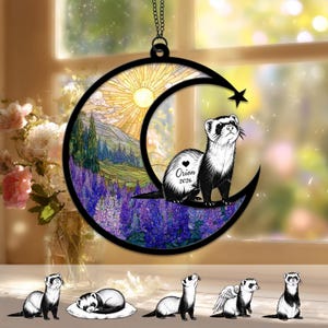 May include: A circular hanging ornament featuring a black crescent moon frame with a star. Inside is a stained-glass style illustration of a ferret, a sun, mountains, and purple flowers. The ferret has a heart, the name Orion, and 2026 printed on it.