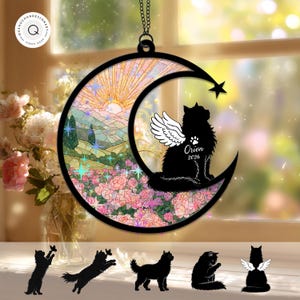 May include: A circular hanging ornament featuring a black crescent moon frame with a stained-glass style landscape of pink flowers and a sunrise. A black cat with white angel wings sits on the moon, with the text Orion 2026 and a paw print.