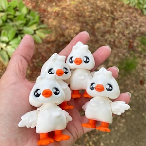 May include: Four white duck figurines with orange feet and beaks, and black eyes. The ducks have small wings and are held in a hand. The background is blurred with green foliage.
