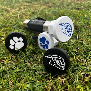 May include: A set of hockey stick plugs and a hockey stick handle, resting on grass. The plugs are white and black, with blue and white graphics of a hockey helmet and a paw print. The handle is wrapped in black tape.