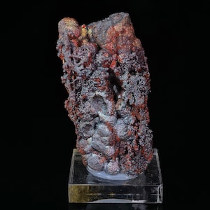 Puede incluir: Un gran espécimen mineral iridiscente, morado y rojo con una superficie rugosa y texturizada. El mineral se muestra en una base de acrílico transparente.