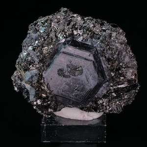Peut inclure: Une formation cristalline hexagonale sombre avec une structure complexe et stratifiée. Le cristal est d'un noir profond et réfléchissant, entouré d'une matrice de fragments minéraux sombres, scintillants et plus petits. Le spécimen repose sur une base noire sur fond noir.