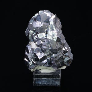 Può includere: Un gruppo di cristalli di colore grigio scuro, metallici e iridescenti su un supporto trasparente. I cristalli hanno una forma geometrica appuntita.