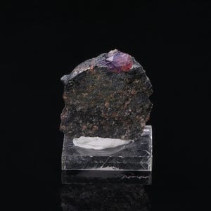 May include: A dark, rough-textured rock with a small, faceted, purple and red crystal on top. The rock sits on a clear, rectangular display stand against a black background. The crystal is the focal point.