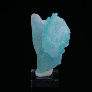 May include: A large, translucent, light blue crystal with a unique, organic shape. The crystal is displayed on a small, white pedestal.