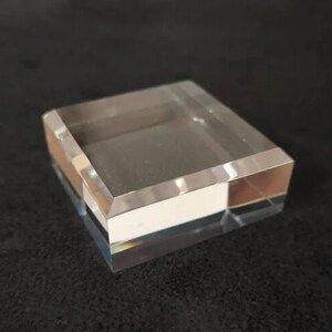 May include: A clear acrylic display stand with a square base and beveled edges. The stand is made of two layers of acrylic, with the top layer being slightly larger than the bottom layer.