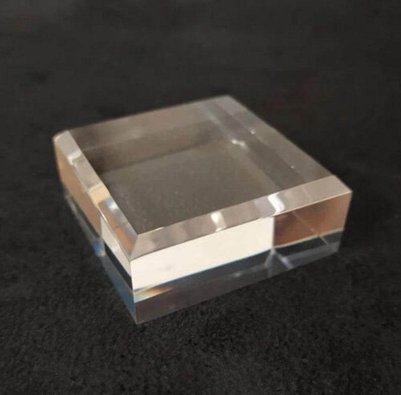 May include: A clear acrylic display stand with a square base and beveled edges. The stand is made of two layers of acrylic, with the top layer being slightly larger than the bottom layer.