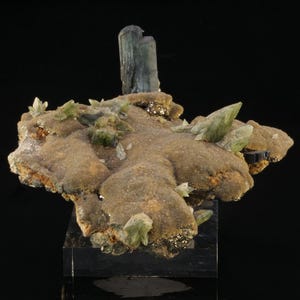 May include: A mineral specimen featuring a large, dark, vertical crystal column rising from a textured, tan-colored matrix. The surface is adorned with smaller, pale green, pointed crystals and clusters of metallic, golden-hued pyrite formations.