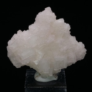 Peut inclure: Un groupe de cristaux d'halite cubiques translucides, rose pâle. Les cristaux sont regroupés en une forme irrégulière et rugueuse. Les cristaux sont translucides et ont une légère teinte rose. Les cristaux sont sur un petit piédestal blanc.