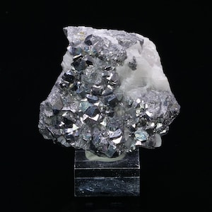 Può includere: Un gruppo di cristalli grigi metallici, di colore argenteo, su una matrice bianca. I cristalli sono appuntiti e hanno una superficie riflettente. Il campione è esposto su un supporto in acrilico trasparente.
