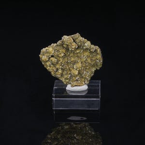 May include: A rough, golden-colored mineral specimen with a textured surface, displayed on a clear acrylic stand. The mineral has a crystalline appearance and sits atop a small, white base.