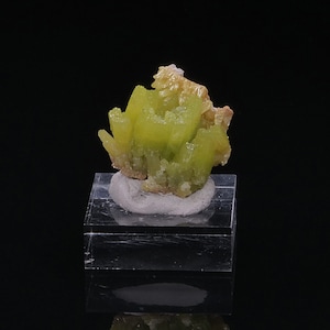 Puede incluir: Un grupo de cristales de color amarillo verdoso brillante sobre una pequeña base blanca. Los cristales están dispuestos en forma de flor.