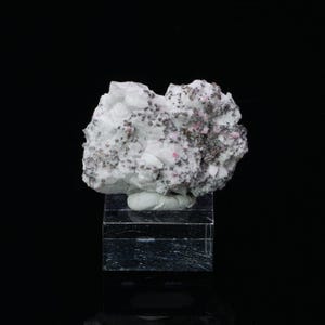 May include: A white mineral specimen with small, dark gray and pink crystals. The specimen is displayed on a clear acrylic base.