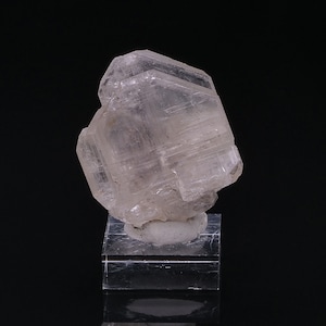 Könnte beinhalten: Ein klares, facettierter, farbloser Kristall-Exemplar auf einem kleinen, klaren Präsentationsständer. Der Kristall hat eine geometrische Form und ist durchscheinend.