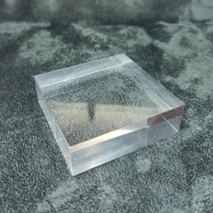 May include: A clear acrylic square display stand. The stand is made of thick, clear acrylic and has a smooth, polished finish.