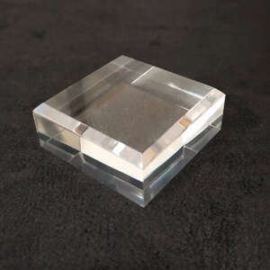 May include: A clear acrylic display stand with a square base and beveled edges. The stand is made of thick, durable acrylic and is perfect for displaying small items.