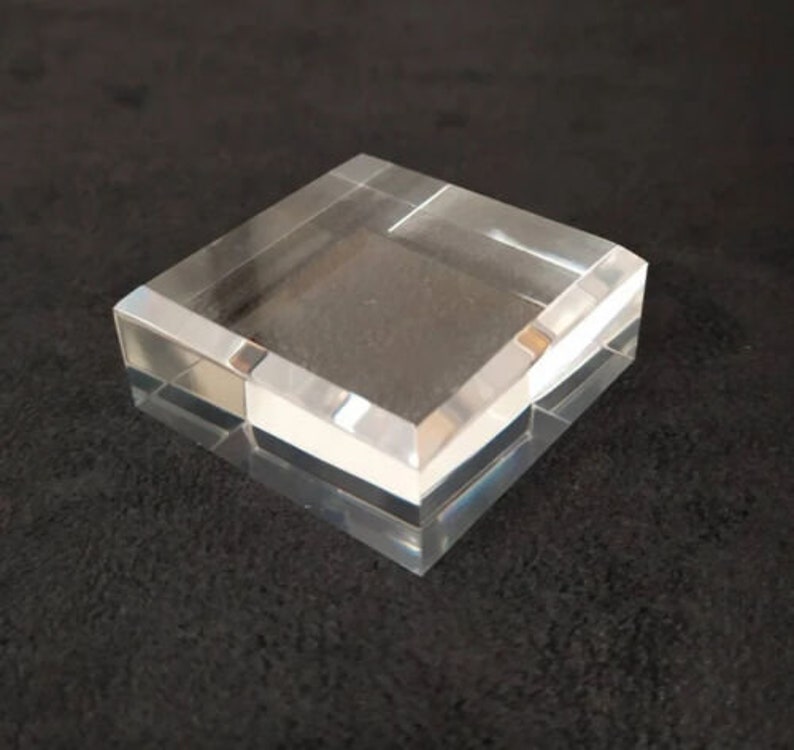 May include: A clear acrylic display stand with a square base and beveled edges. The stand is made of thick, durable acrylic and is perfect for displaying small items.